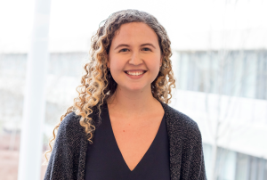 Headshot of Natasha Vitkin in a black shirt, grey cardigan. She has curly blonde hair and is smiling at the camera. Out of focus in the background is a white building and some deciduous trees.