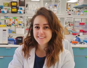 Headshot of Sarah Nersesian in a lab coat, black shirt, disposable gloves. Behind Sarah is a shelf full of lab equipment, including Erlenmeyer flasks