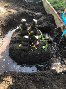 Four tiny snowmen made out of mud. They are on a mud island, with a blue watering can pouring water into a moat around them. The mud people are wearing hats made out of zucchini.